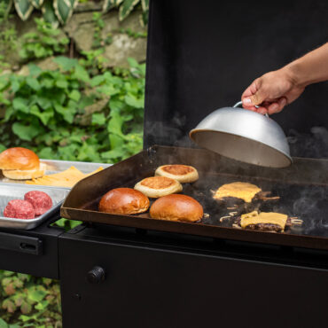Outdoor image of cheese melting dome over a grill with burger patties and buns